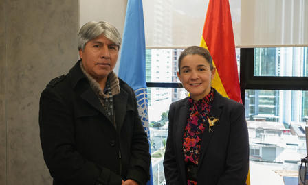 Two adults stand in a modern office with large windows; a red-and-yellow flag behind them.