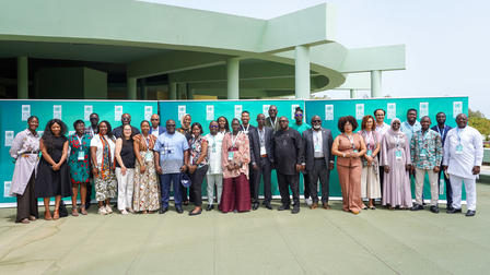Photograph of a group of people standing in front of a green building on a sunny day.