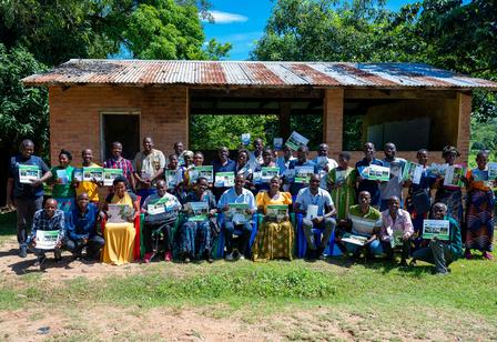 Group of people, including several in wheelchairs, pose outside a rustic building on a sunny day.