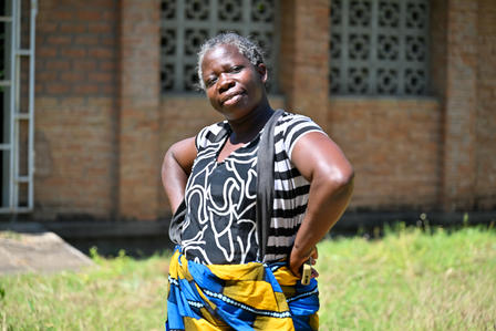 Man outdoors before brick building, hands on hips, wearing patterned sleeveless shirt and wrap.