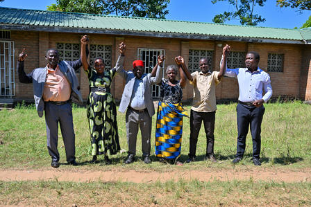 Group of people standing in a line outside a brick building with a blue roof, arms raised.