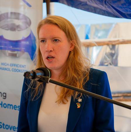 Photograph of a blonde woman in a blue blazer speaking into a microphone at a podium.