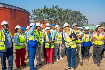 Group of construction workers in safety vests and hard hats listening to a guide on a sunny site.