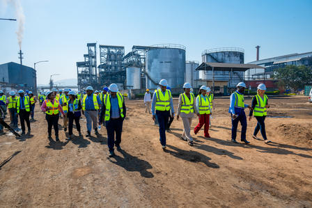 Group of construction workers in yellow high-visibility vests walking on a dirt site with buildings in the background.