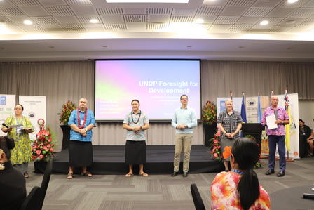 Group of people on stage at a conference ceremony, framed by potted plants.
