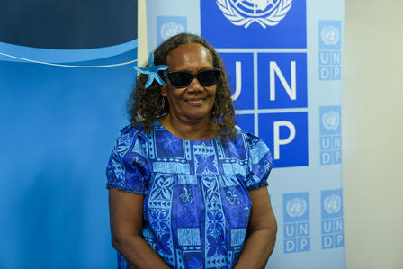 Smiling woman in a blue patterned dress and sunglasses stands before UNDP backdrop.