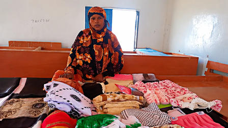 A woman in a colorful floral hijab stands beside a bed covered with folded clothes in a small room.