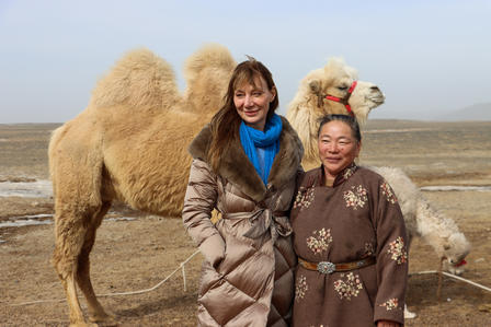 Two women in coats stand in a desert with two camels behind them.