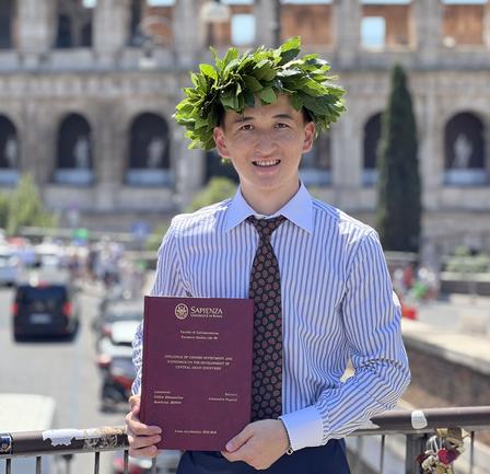 Graduate wearing a laurel wreath, holding a purple diploma outdoors.
