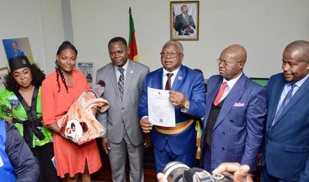 Group of officials in formal attire posing with a document in a room with a Nigerian flag.