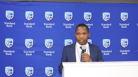 Man in suit speaks at a podium in front of a blue backdrop featuring shield logos.