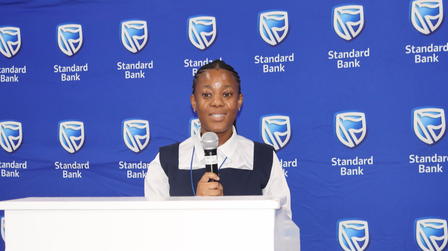 Photograph of a woman at a podium speaking, blue backdrop with Standard Bank logos.