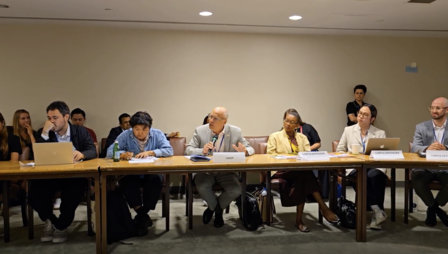 Photograph shows a group of people seated at a long conference table in a meeting room, with papers in front.