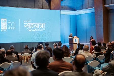 Speaker at podium addressing an audience in a conference hall; blue backdrop with a 60 years logo.
