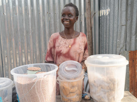 Smiling woman at a market stall with clear containers and a corrugated metal backdrop.