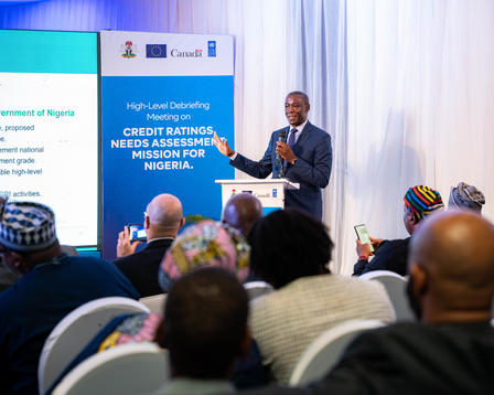 Photograph of a man in a suit speaking at a conference podium, with blue banners and audience.