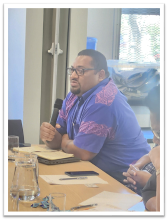 Man in a purple-patterned shirt with glasses speaks into a microphone at a conference table.