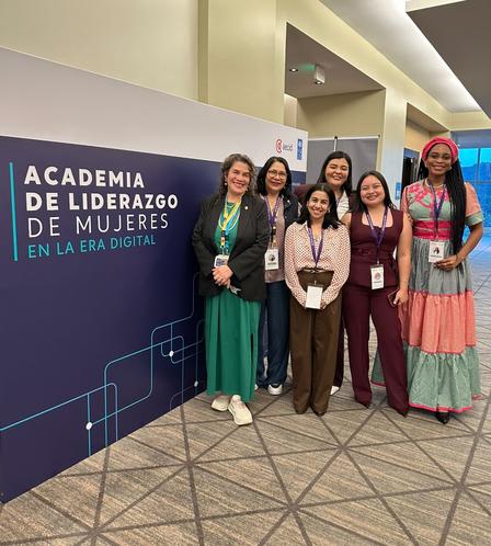 Five women with conference badges pose beside a banner reading Academia de Liderazgo de las Mujeres.