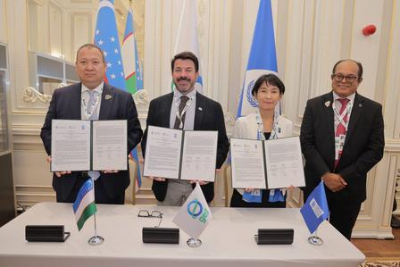 Four suited officials hold documents at a formal signing ceremony; flags in an ornate room behind.