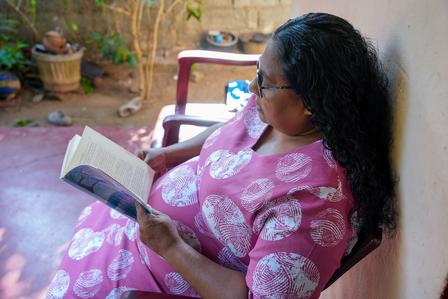 Woman in a pink patterned dress reads a book while seated on a chair outdoors.