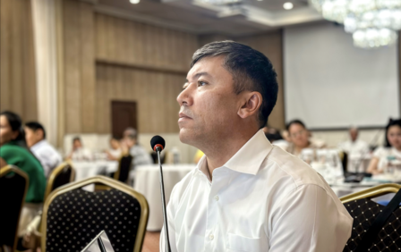 Photo: man in a white shirt seated at a conference, looking ahead; crowd in background.