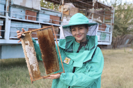 Smiling beekeeper in teal protective suit holding a honeycomb frame outdoors.