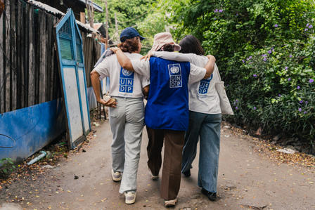 Three friends walk arm-in-arm down a dirt path, with trees and a wooden fence beside them.