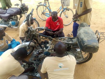 Group of men repair a motorcycle on dusty ground; engine exposed.