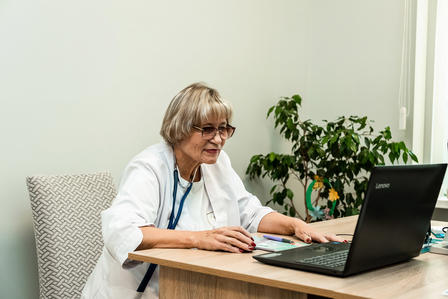 A woman healthcare professional wearing a white medical coat and a stethoscope sits at a desk, using a laptop during an online consultation or administrative task in a bright, modern clinic office. A potted plant is visible in the background.