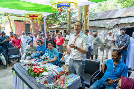 Photograph of an outdoor award ceremony with a man speaking at a table, flowers and audience.