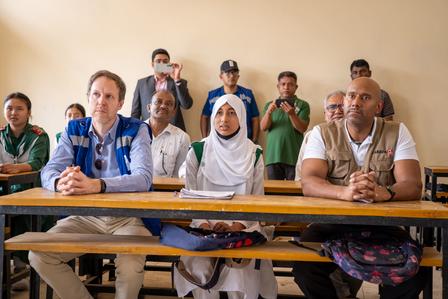 Diverse group of people seated at wooden desks in a classroom, listening attentively.