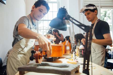 Two luthiers in a sunlit workshop shape string instruments at a workbench.