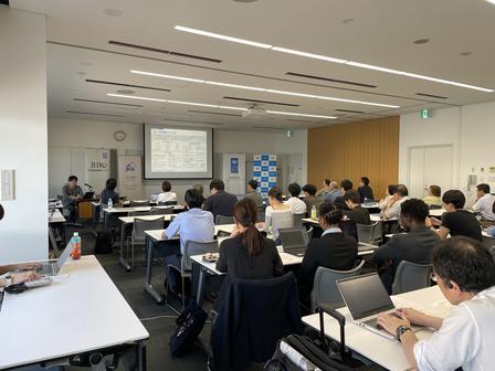 Lecture hall with students seated at desks, a presenter at the front near a projector screen.