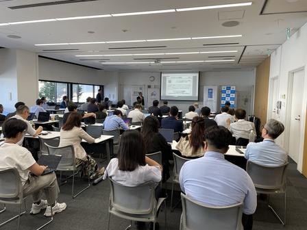 Audience seated in a bright conference room watches a slide presentation at the front.