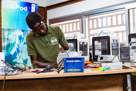 Man in a green shirt leaning over a cluttered workshop bench with tools and electronics.