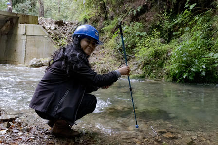 Photograph of a person wearing a blue helmet kneeling by a shallow stream, holding a long pole.