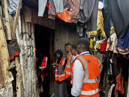 People in orange safety vests in a narrow alley between shanties with clothes hanging overhead.