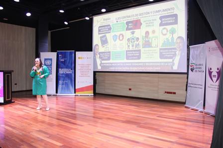 Presenter in green on stage with a colorful infographic projected on screen.