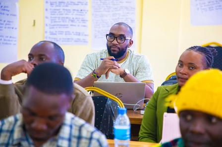 Diverse adults in a classroom; bearded man with glasses at center, hands clasped.