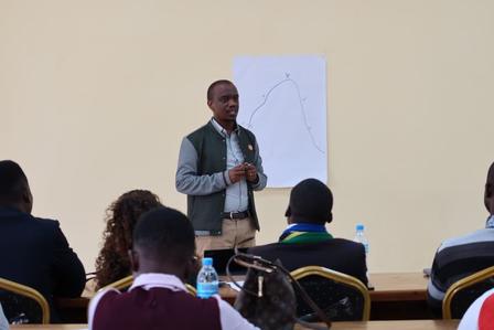 Male presenter in a vest speaks at the front of a classroom, with a whiteboard behind.