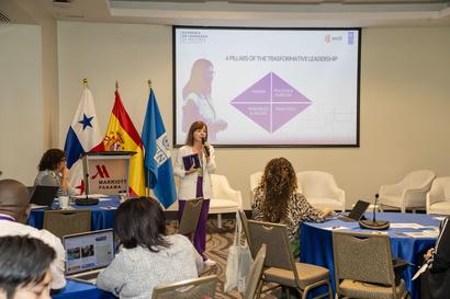 Photograph: female presenter at podium in conference room; purple diamond slide on projector.