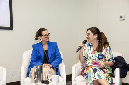 Two women sit on a panel; one in blue blazer, the other in floral dress speaking into a microphone.
