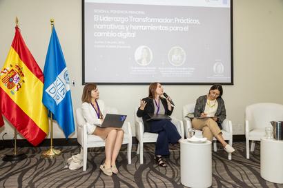 Panel discussion with three women seated on stage, flags to the left and a projected screen behind.