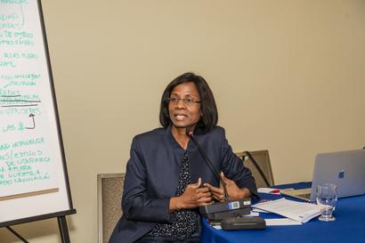 Photograph of a smiling woman in a dark blazer presenting at a table with a blue tablecloth and whiteboard.