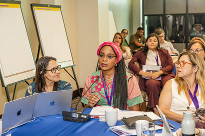 Photograph of diverse women at a workshop, seated around a blue table with laptops.