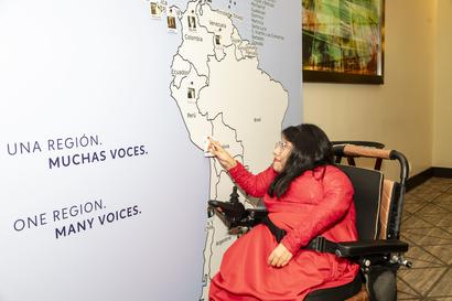 Photograph of a woman in a red dress in a wheelchair signing beside a map banner.