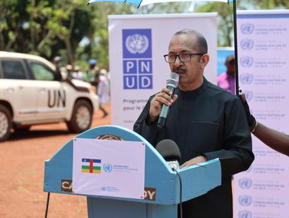 Man speaks at podium during UNDP event; banners and a UN vehicle in background.
