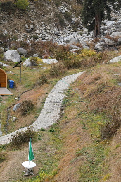 Small orange dome tent on a hillside campsite with a winding stone path and a blue trash bin nearby.
