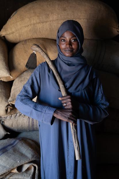 Woman in blue hijab stands beside stacked burlap sacks in a dim room.