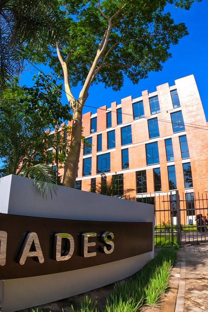 Modern brick building behind a curved stone sign reading MADES, with trees and blue sky.